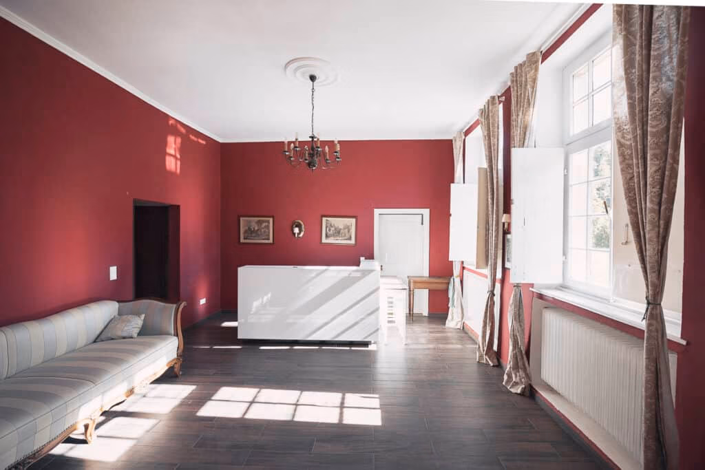 Raum mit roten Wänden, großen Fenstern, gestreiftem Sofa, Kronleuchter und weißem Empfangstresen – ideal für Hochzeiten oder Firmenevents.