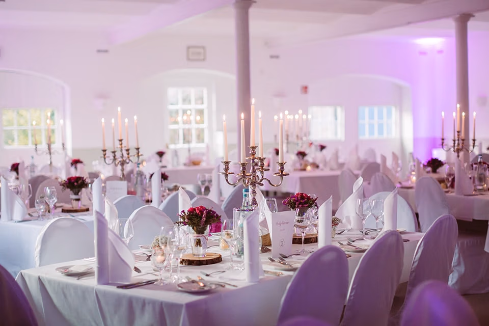 Eleganter Bankettsaal f&uuml;r Firmenveranstaltungen mit wei&szlig;em Tischgedeck und violetten Blumendekorationen.