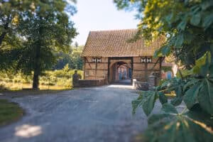 Firmeneventlocation 14 Gebäude aus Backstein und Holz mit Torbogen, perfekt als Hochzeitslocation neben einer von Bäumen gesäumten Schotterstraße.